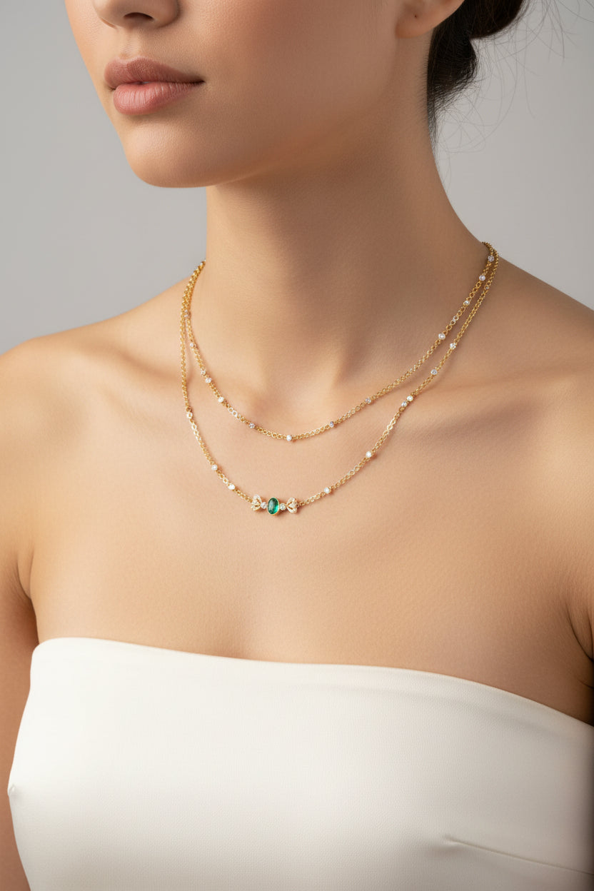 A delicate double-strand gold necklace with small clear stones and a central bow-shaped charm featuring light blue and yellow accents.